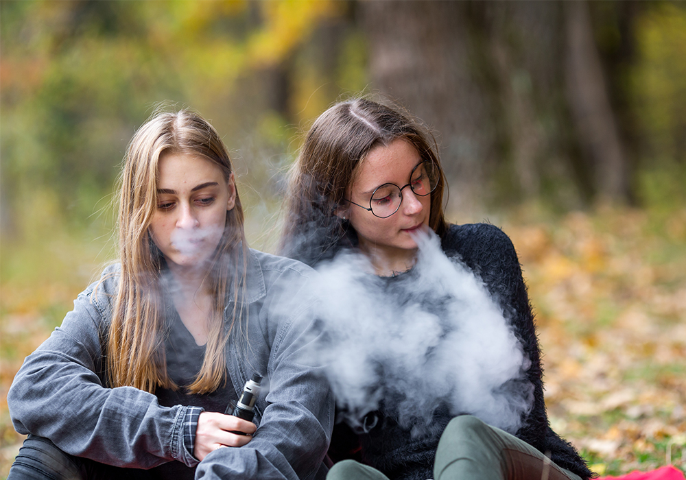 Two women are sitting down while smoking e-cigarettes