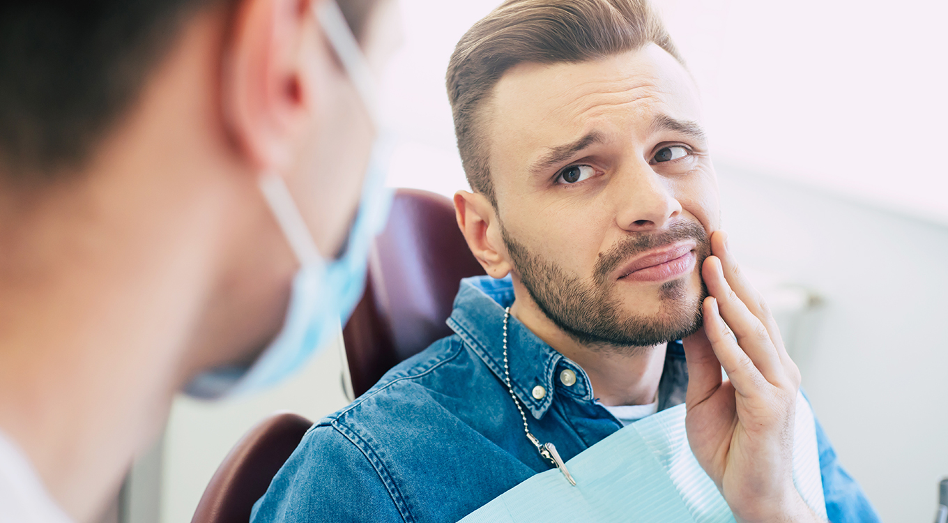 A man sitting in a dentist chair and touching his jaw while looking at the dentist